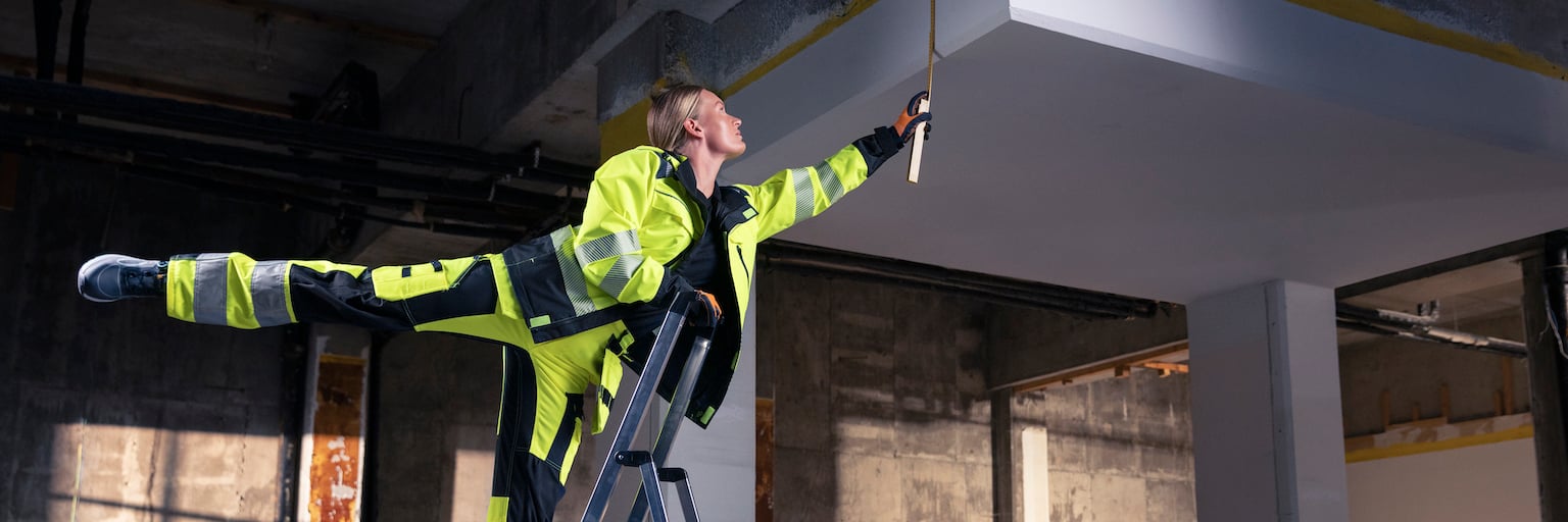 Kvinne i synlighetsklær står høyt på en stige med det ene beinet strukket rett bakover og måler takhøyden med et målebånd.