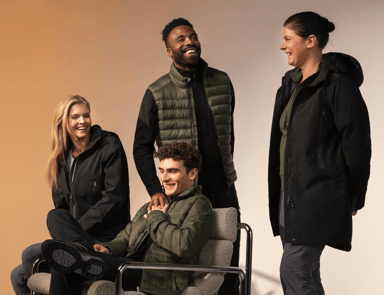 Four colleagues laugh together in work clothes from the South West – warm jackets, vests and functional trousers in black and olive green. Clickable link, read more about Båstadgruppen's sustainability work.