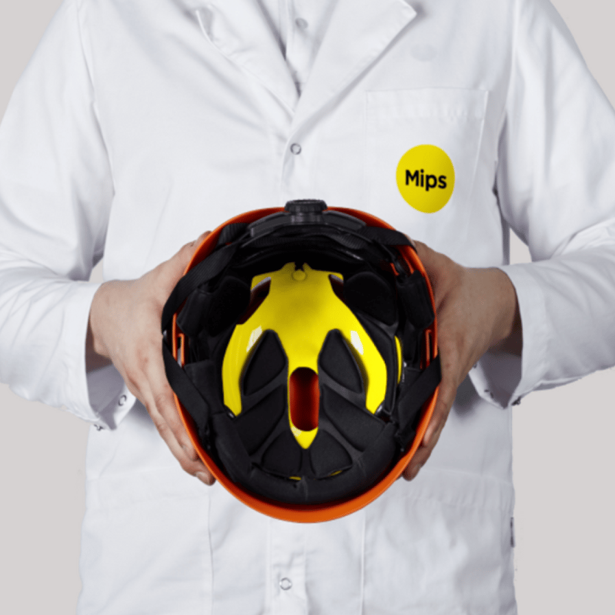 Eine Person in einem weißen Laborkittel mit dem Mips-Logo hält einen Schutzhelm mit einem gelben Mips-System in der Hand